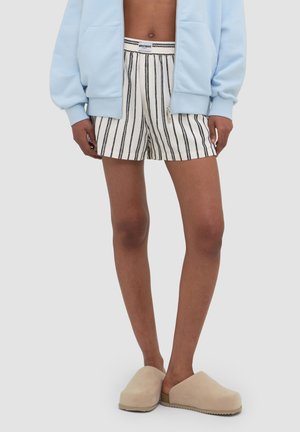 Person iført en lyseblå jakke, hvide shorts med sorte lodrette striber og beige slip-on sko, stående mod en ensfarvet baggrund.