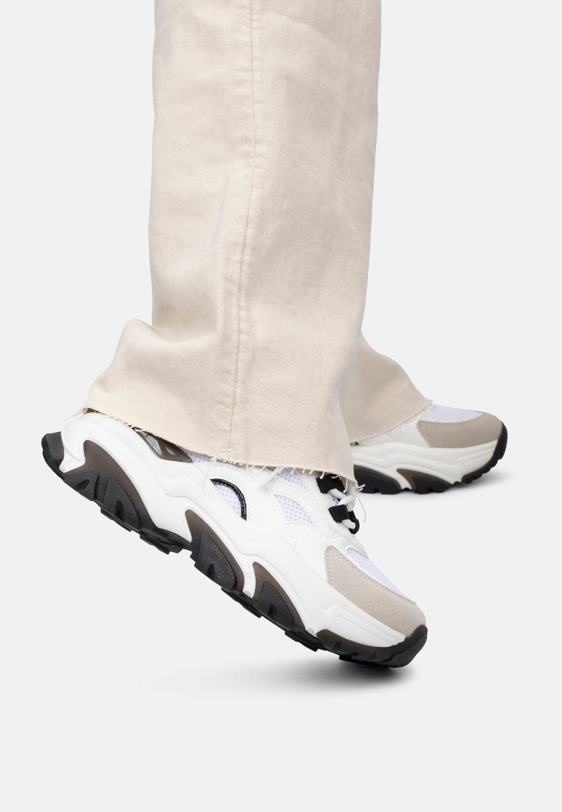 Baskets blanches épaisses avec des accents beiges et noirs, des panneaux en maille et des semelles texturées, associées à un pantalon large de couleur claire.