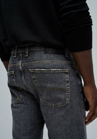 Jeans de mezclilla gris oscuro con un corte ajustado. Las características incluyen dos bolsillos traseros, costuras naranjas y una etiqueta de cuero negro en la cinturilla.