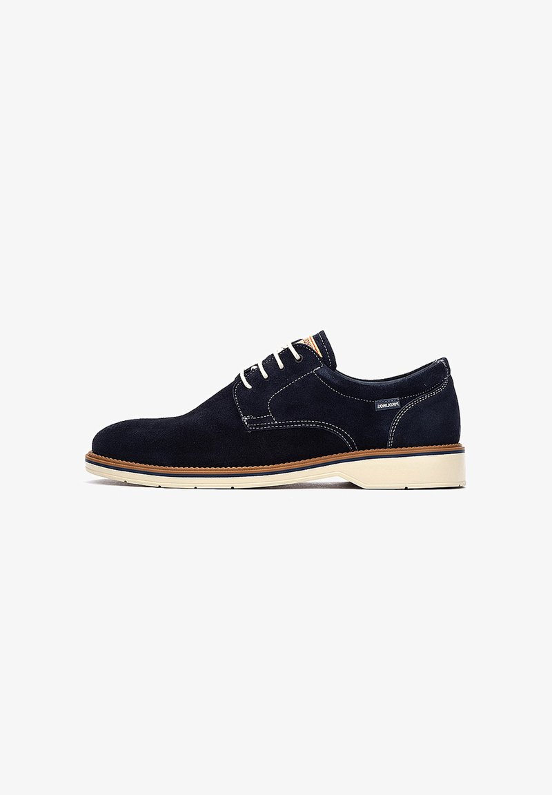 Zapato de hombre azul marino de ante con cordones, suela blanca y ribete marrón, mostrado de perfil sobre fondo blanco.