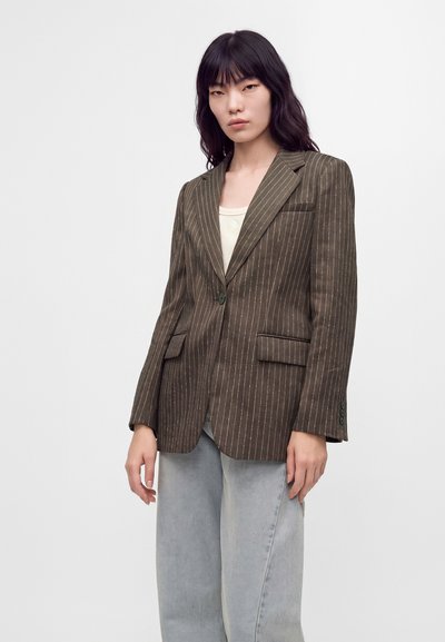 Femme portant un blazer à fines rayures marron sur un haut crème et un jean large gris clair, debout devant un fond clair uni.