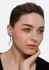 Orecchini in metallo nero con un design a spirale attorcigliata, indossati all'orecchio sinistro. Il soggetto ha la pelle liscia e sta toccando il mento.