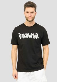 Uomo con capelli corti e scuri e barba indossa una maglietta nera con il testo bianco "Disclaimer", abbinata a pantaloni bianchi, su uno sfondo grigio semplice.