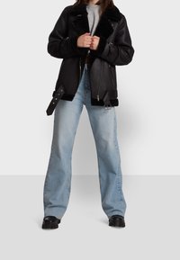 Svart läderjacka med konstpälskrage och manschetter, dubbel dragkedjeslutning och justerbara remmar, parat med ljusblå vida jeans.