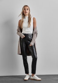 Zwarte leren broek gecombineerd met een wit geribbeld mouwloos topje en een lange beige cardigan. Witte sneakers met een gumrubberen zool maken de outfit compleet.