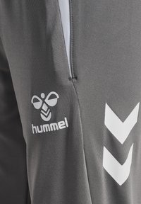 Close-up van grijze sportbroek met wit hummel-bijlogo en dubbele chevronpatroon bij de ritszak.