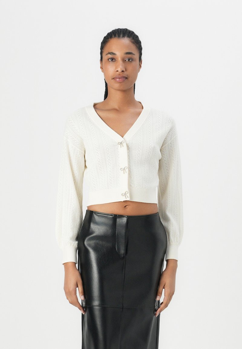Cardigan crème coupé avec un col en V, présentant un design en tricot texturé et des boutons en forme de nœud décoratifs, assorti à une jupe crayon noire.