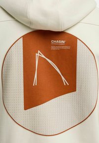 Creme kleurige hoodie met een groot oranje cirkelontwerp, voorzien van witte geometrische patronen en het merk CHASIN'.