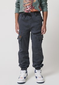 Cargo hlače iz temno sive tkanine, z bočnimi žepi, elastičnimi zapestji in pasom z vrvico. V kombinaciji z belimi in mornarsko modrimi supergami.