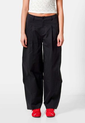 Frau trägt hoch taillierte, weit geschnittene schwarze Hosen, weißes, kurzes Spitzenoberteil und rote Puma-Sneaker, steht vor einem schlichten weißen Hintergrund.