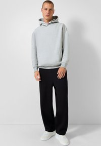 Giovane uomo in piedi che indossa un hoodie grigio chiaro, pantaloni neri larghi e sneakers bianche, su uno sfondo grigio chiaro uniforme.
