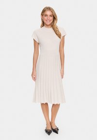 Light beige ribbed knit dress with short sleeves, fitted bodice, and a pleated skirt. Worn with black pointed toe heels.