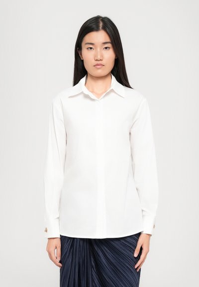 Chemise blanche à manches longues en tissu lisse, avec un col classique, une fermeture à boutons et des accents dorés sur les poignets.
