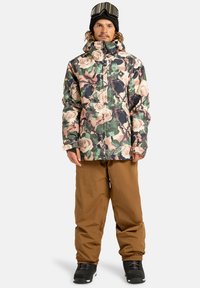 Mężczyzna w wielokolorowej kurtce w kwiatowy wzór, brązowych spodniach narciarskich, czarnych butach, czarnej czapce beanie i narciarskich goglach na czole.