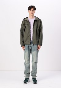 Grønn hettejakke med glidelås, frontlommer, brukt over en lys lilla skjorte. Blå jeans og grønne sneakers fullfører antrekket.
