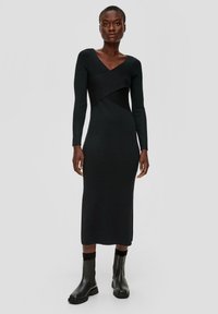 Schwarzes langärmliges Kleid mit V-Ausschnitt, das einen taillierten Oberkörper und ein asymmetrisches Design aufweist. Kombiniert mit schwarzen Stiefeletten. Glatte Textur.