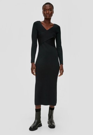 Schwarzes langärmliges Kleid mit V-Ausschnitt, das einen taillierten Oberkörper und ein asymmetrisches Design aufweist. Kombiniert mit schwarzen Stiefeletten. Glatte Textur.