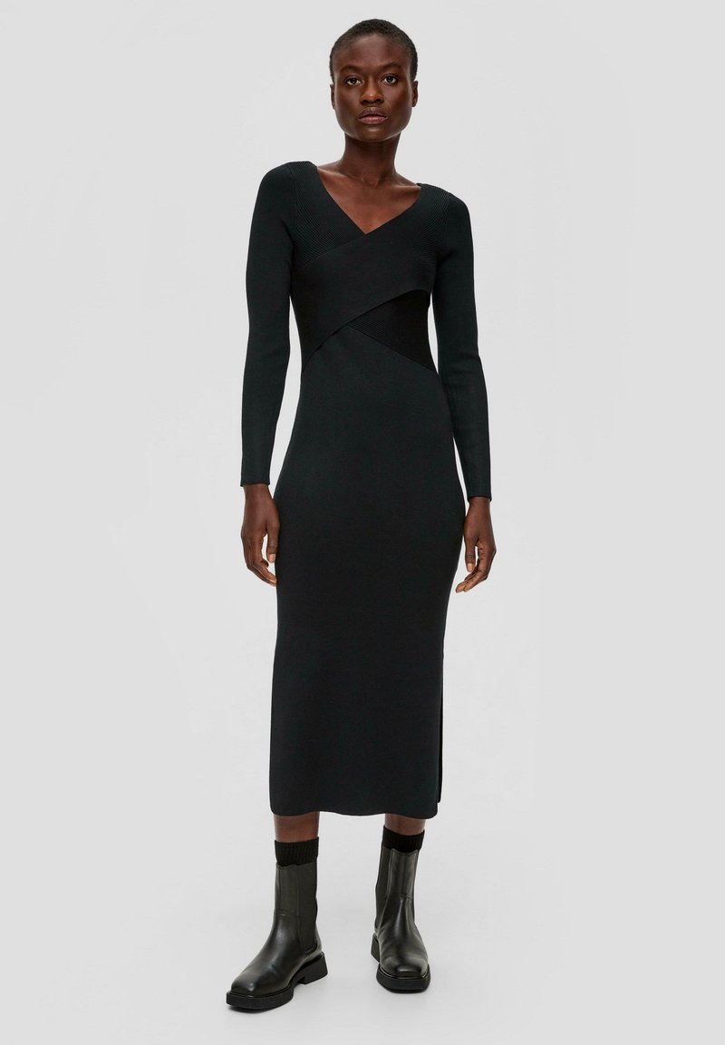 Schwarzes langärmliges Kleid mit V-Ausschnitt, das einen taillierten Oberkörper und ein asymmetrisches Design aufweist. Kombiniert mit schwarzen Stiefeletten. Glatte Textur.