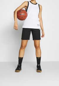 Biała koszulka na ramiączkach Nike z czarnym wykończeniem, czarne szorty, czarne skarpetki w wzory i czarne sneakersy, trzymająca koszykówkę, prezentująca odzież sportową.