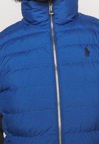 Chaleco acolchado azul con frente con cremallera y alto cuello; presenta costuras en alfileres horizontales y un pequeño logo en el pecho.