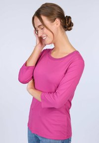 Fuchsia Langarmshirt mit rundem Hals und zarter Stickerei, kombiniert mit hellblauen Jeans, getragen von einer Person vor einem neutralen Hintergrund.