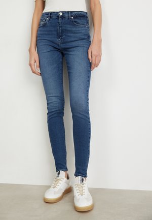 Model iført slim-fit blå jeans og hvide sneakers med beige såler, stående op ad en ensfarvet hvid væg og neutral gulv.