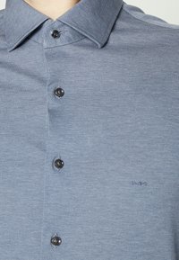 Lichtblauw overhemd met knopen en een puntige kraag, voorzien van een klein geborduurd 'MK' logo op de linkerborst.