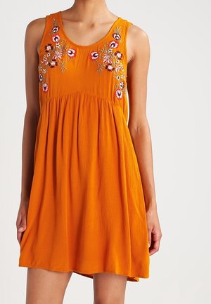Ærmeløs orange kjole med indsnævret talje og blomsterbroderi på brystet, båret af en person med armene afslappet ned langs siden.