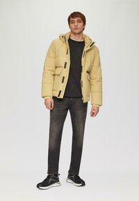 Pufferjack in lichtgeel met een ritssluiting aan de voorkant, zwarte accenten en meerdere zakken. Gecombineerd met donkere jeans en zwarte sneakers.