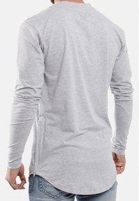 Helles graues Langarmshirt aus leichtem Stoff, mit Rundhalsausschnitt, seitlichem Reißverschluss und abgerundetem Saum. Dezentes Logo in der Nähe des Kragens.