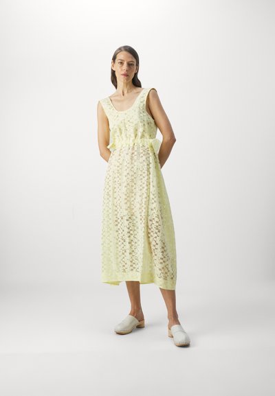 Robe midi en dentelle de couleur jaune doux avec une coupe ample, un décolleté rond et une ceinture à la taille. Motif texturé sur toute la surface, associée à des sabots blancs.