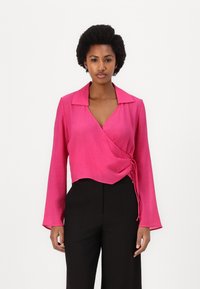Pink wrap blouse with long sleeves, featuring a collared neckline, textured fabric, and a side tie detail. Worn with black high-waisted pants.