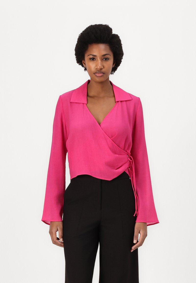 Pink wrap blouse with long sleeves, featuring a collared neckline, textured fabric, and a side tie detail. Worn with black high-waisted pants.
