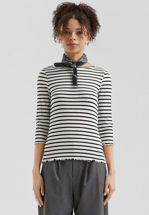 Gestreepte ribbeltop in zwart en wit met driekwart mouwen en een gerimpelde zoom. Gestyled met een zwarte sjaal om de nek.