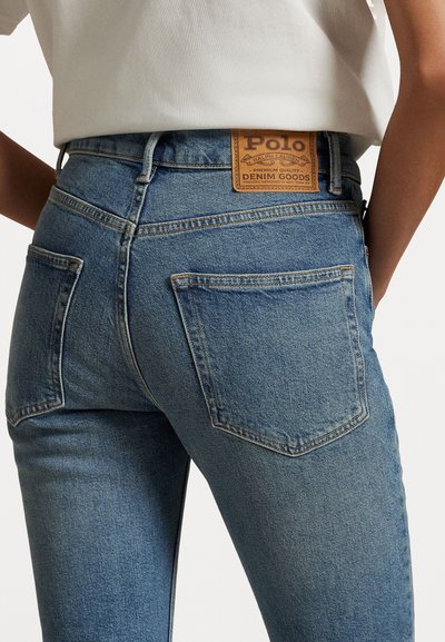 Close-up de uma pessoa a usar jeans de ganga azuis com bolsos traseiros e uma etiqueta de pele castanha da Polo Ralph Lauren na faixa da cintura.