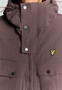 Lila jacka med krinklad textur, silverfärgade tryckknappar, justerbar huva och två framfickor. Har en broderad gul örnlogotyp.