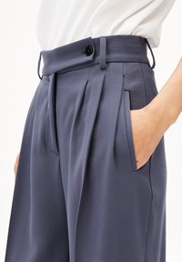 Marineblaue, gefaltete Hose mit flachem Bund, einem Knopf und seitlichen Taschen, gefertigt aus einem glatten Stoff mit einer subtilen Textur.