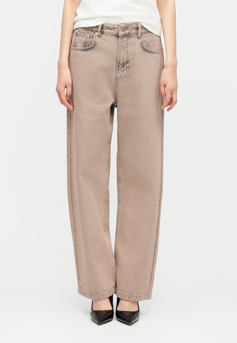 Μπεζ τζιν με φαρδιά παντελόνια, φορεμένα με μαύρες γόβες με μυτερή μύτη και λευκό τοπ που έχει μπει μέσα στο παντελόνι, εμφανιζόμενα από τη μέση μέχρι τα πόδια.