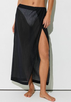 Femme portant une jupe noire semi-transparente avec une fente haute sur la cuisse, dévoilant des sous-vêtements noirs et des pieds nus, devant un mur blanc.
