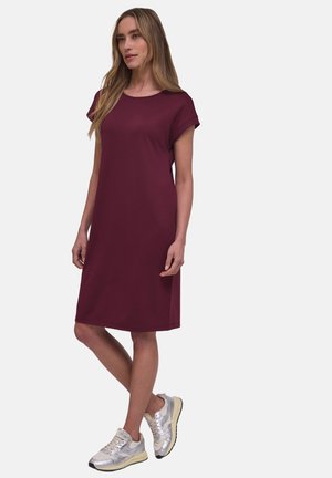 Donna in piedi con le gambe incrociate, indossa un vestito bordeaux a maniche corte fino al ginocchio e sneakers argento su uno sfondo semplice.