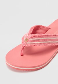 Roosa kummist flip-flop, millel on tekstureeritud rihm, millel on valged triibud ja kuldne detail. Lame tallaga, millel on lainekujuline äär.