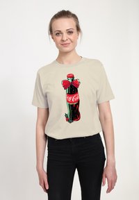 Béžové tričko s grafickým motivem láhve Coca-Cola s červenou mašlí, akcenty cesmíny a živými barvami na světlém pozadí.