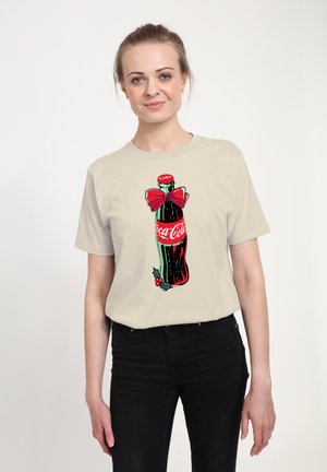 Beige t-shirt met een afbeelding van een Coca-Cola-fles met een rode strik, hulstaccenten en levendige kleuren op een lichte achtergrond.
