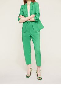 Zelený střihový blazer a odpovídající zkrácené kalhoty z hladké tkaniny, doplněné zelenými sandály na vysokém podpatku se šněrováním.