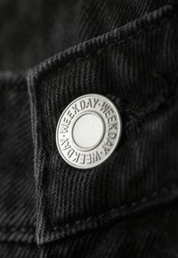 Buton metalic cu textul „WEEKDAY” gravat în jurul marginii, pe un material negru texturat, probabil parte a unui articol vestimentar.