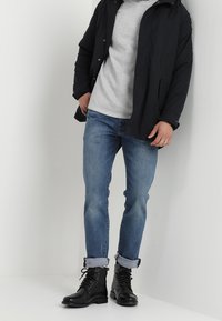 Veste matelassée noire portée sur un pull gris, jean en denim bleu retroussé aux chevilles, et bottines noires à lacets.
