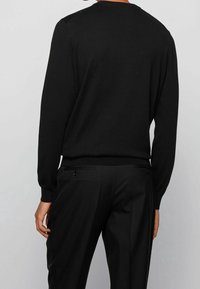 Schwarzer Strickpullover mit langen Ärmeln und rundem Halsausschnitt, kombiniert mit maßgeschneiderten schwarzen Hosen mit zwei Gesäßtaschen.