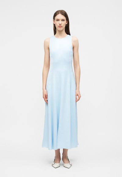 Femme debout, de face, portant une robe maxi sans manches bleu clair et des chaussures blanches pointues, sur un fond blanc uni.