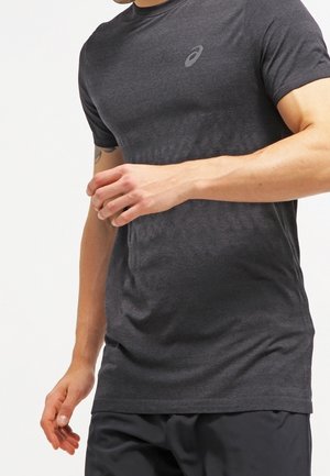 Mężczyzna ubrany w dopasowaną, ciemnoszarą sportową koszulkę z logo Asics i czarne spodenki na jednolitym białym tle.