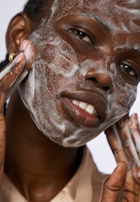 Une personne à la peau foncée applique un nettoyant moussant sur son visage. L'arrière-plan est clair, mettant en valeur la texture des bulles et la douceur de la peau.